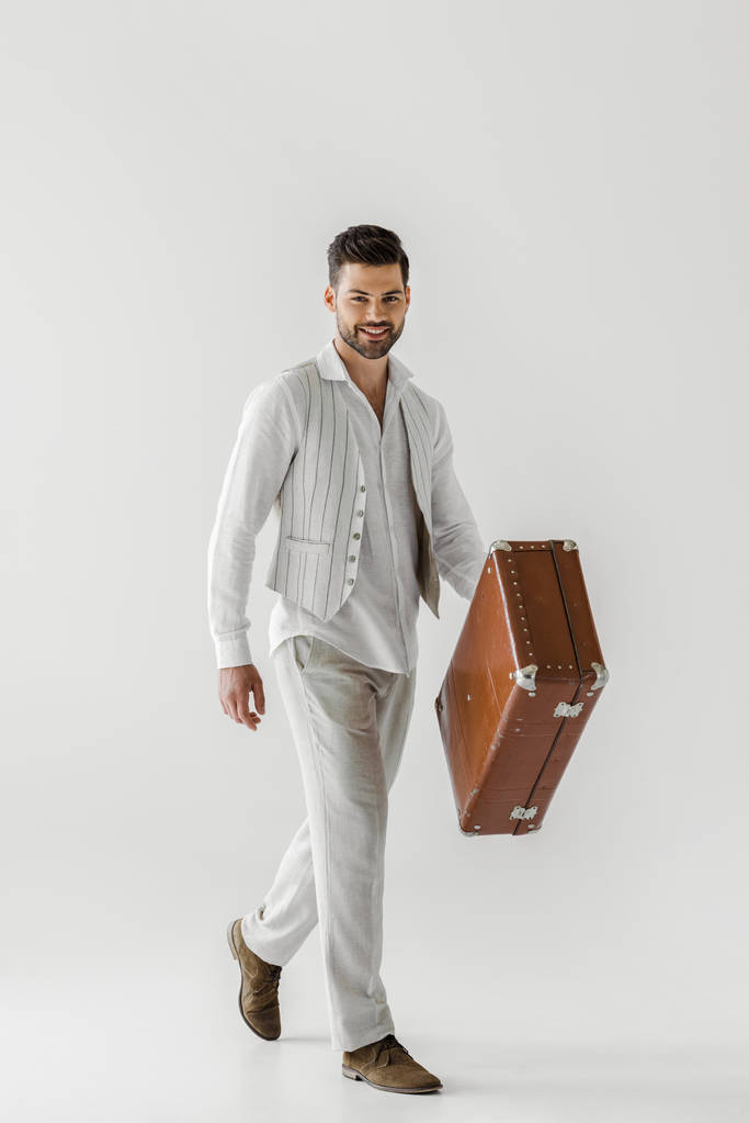灰色の背景に分離されたヴィンテージのスーツケースを運ぶリネンの服の流行の男性旅行者の笑顔 - 写真・画像