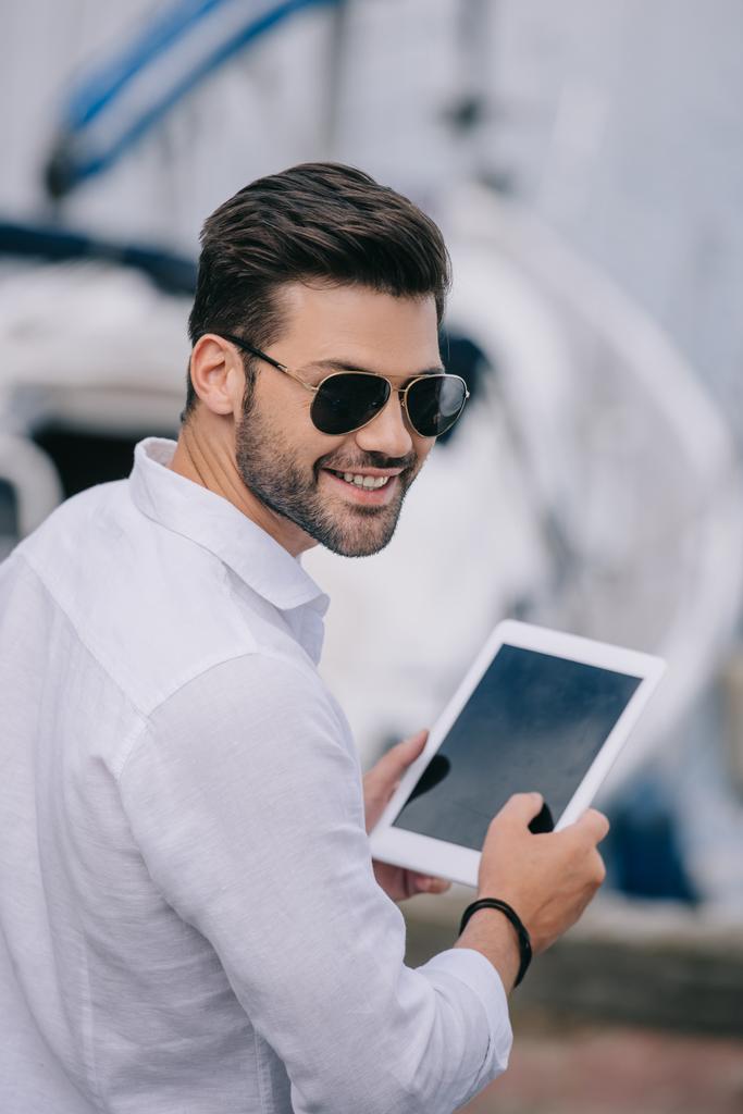όμορφος νεαρός άνδρας με γυαλιά ηλίου που χαμογελάει κάμερα ενώ χρησιμοποιείτε ψηφιακό tablet με την κενή οθόνη - Φωτογραφία, εικόνα