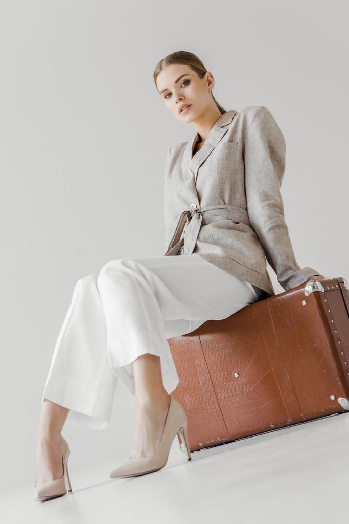 Low-Winkelansicht von stilvollen Frau in Leinenjacke sitzt auf Vintage-Koffer isoliert auf grauem Hintergrund  - Foto, Bild