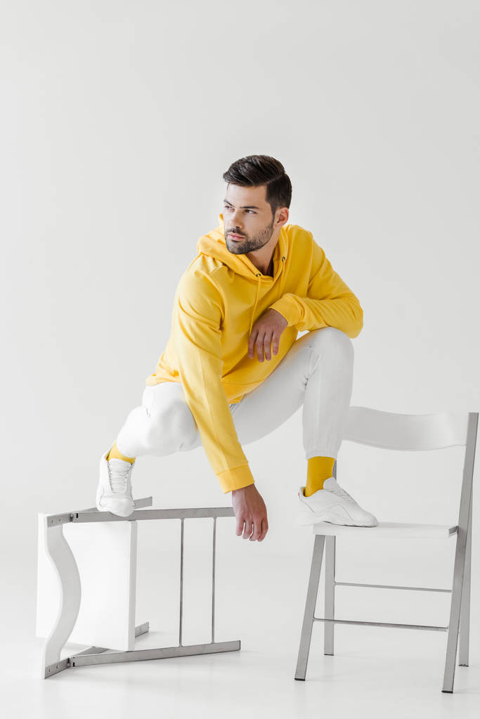 стильний молодий чоловік в жовтому светрі сидить присідає на перевернутих стільцях на білому
 - Фото, зображення
