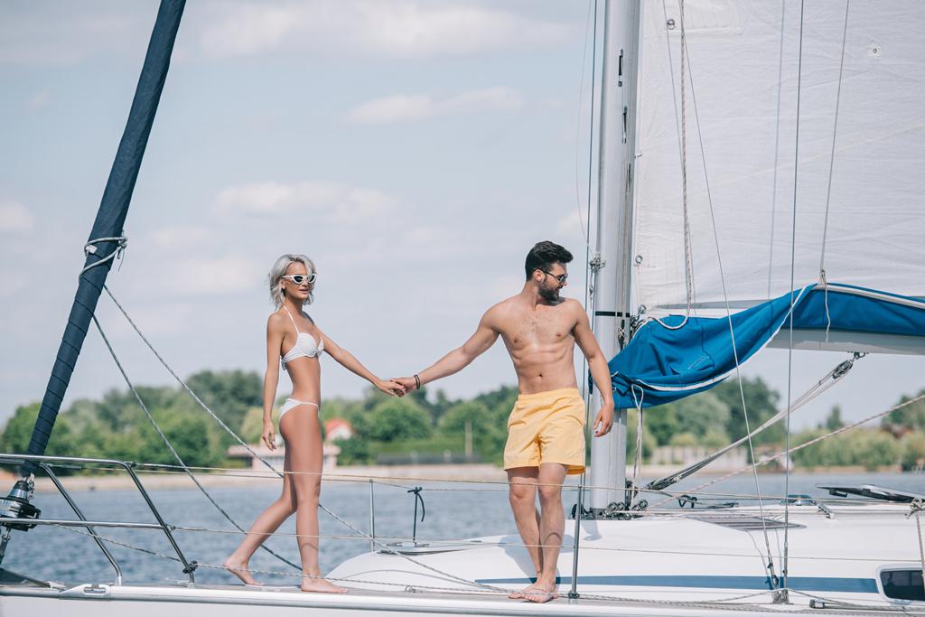 beautiful young couple in swimwear and sunglasses holding hands while spending time together on yacht - Photo, Image