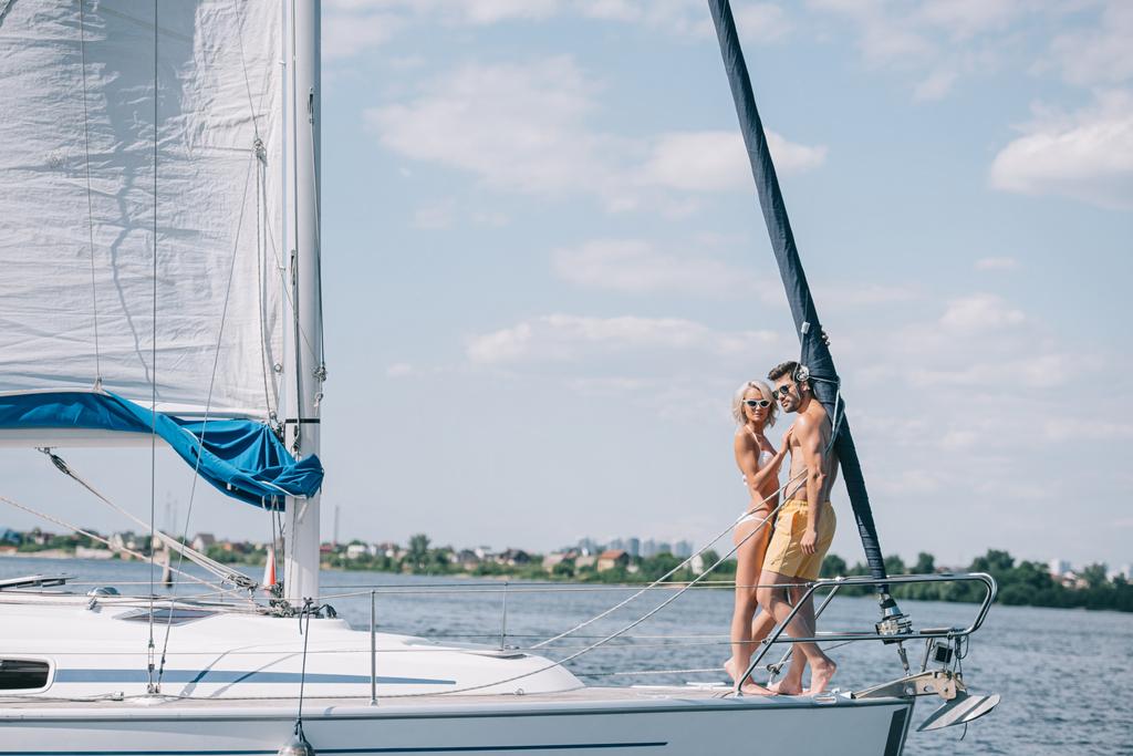 vista completa di bella giovane coppia in costume da bagno e occhiali da sole in piedi insieme su yacht
 - Foto, immagini