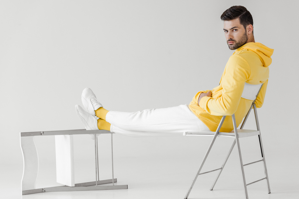 όμορφος νεαρός άνδρας στο κίτρινο hoodie κάθεται χαλαρός στον λευκό και βλέπουν τα φωτογραφικών μηχανών - Φωτογραφία, εικόνα