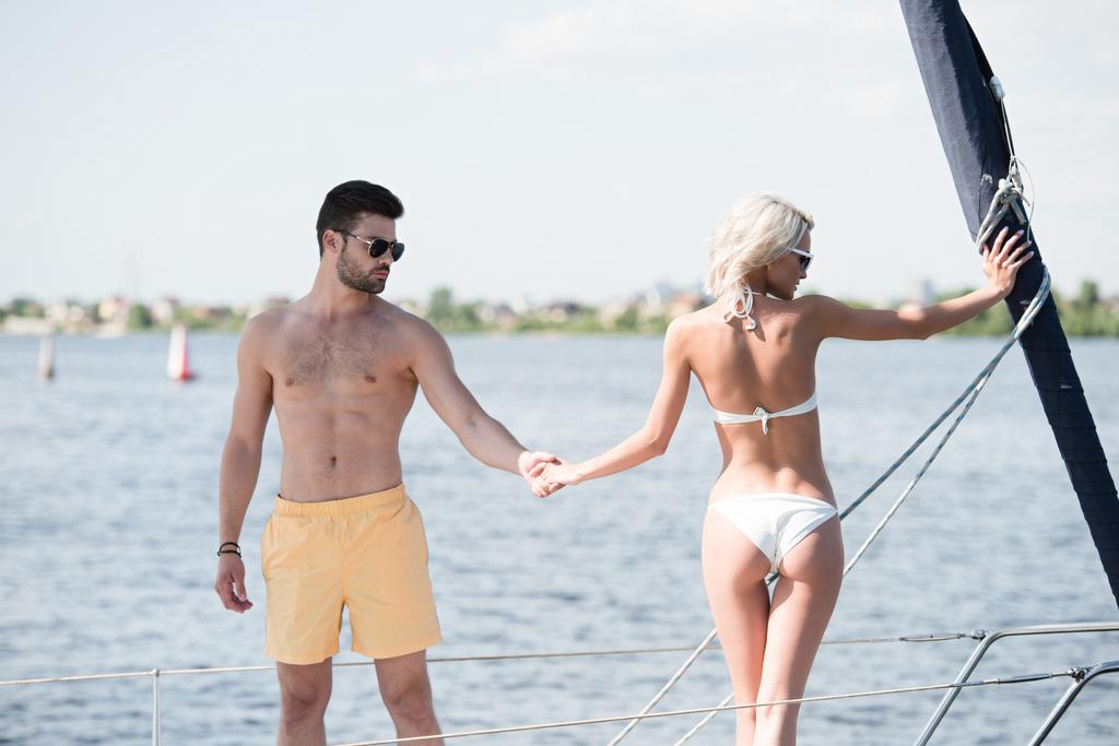 молодая пара в купальниках и солнцезащитных очках держатся за руки, стоя вместе на яхте
 - Фото, изображение