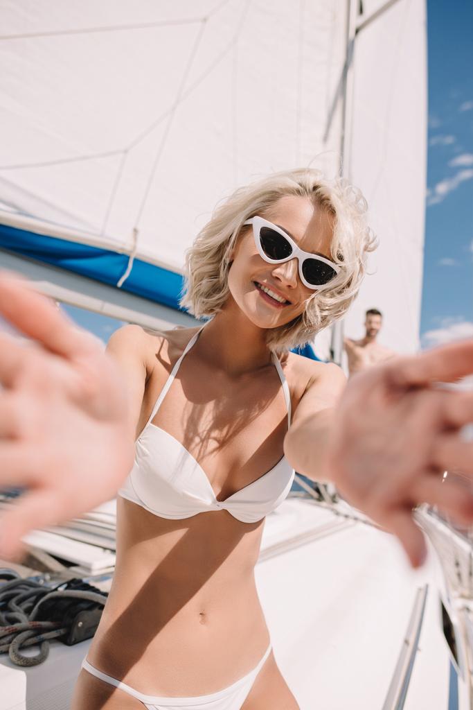 beautiful girl in swimwear and sunglasses reaching hands and smiling at camera on yacht - Photo, Image
