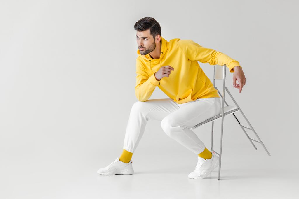 κομψό νεαρό άνδρα σε κίτρινο hoodie κάθεται σε καρέκλα σε λευκό - Φωτογραφία, εικόνα