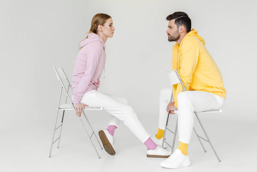 вид сбоку на молодых моделей мужского и женского пола в розовых и желтых капюшонах, сидящих на стульях и смотрящих друг на друга на белом
 - Фото, изображение
