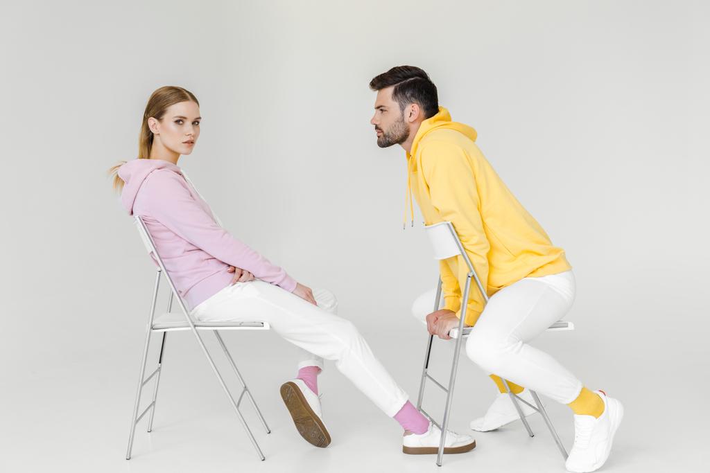 Seitenansicht junger männlicher und weiblicher Models in rosa und gelben Kapuzenpullis, die auf Stühlen auf weißen Stühlen sitzen - Foto, Bild
