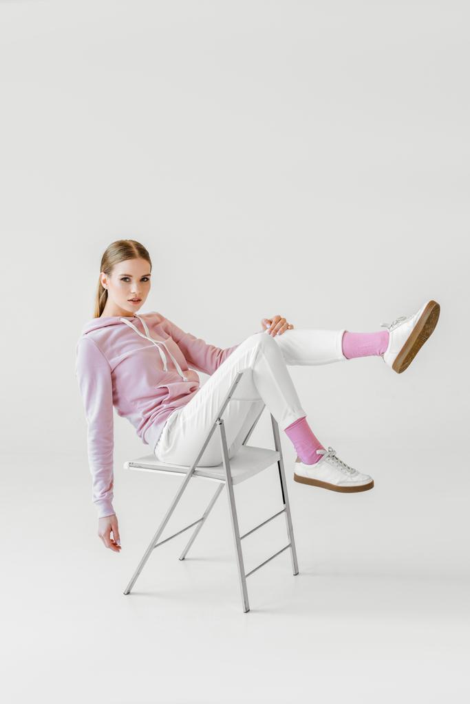 modische junge Frau in rosa Kapuzenpulli sitzt auf Stuhl auf weiß und blickt in die Kamera - Foto, Bild