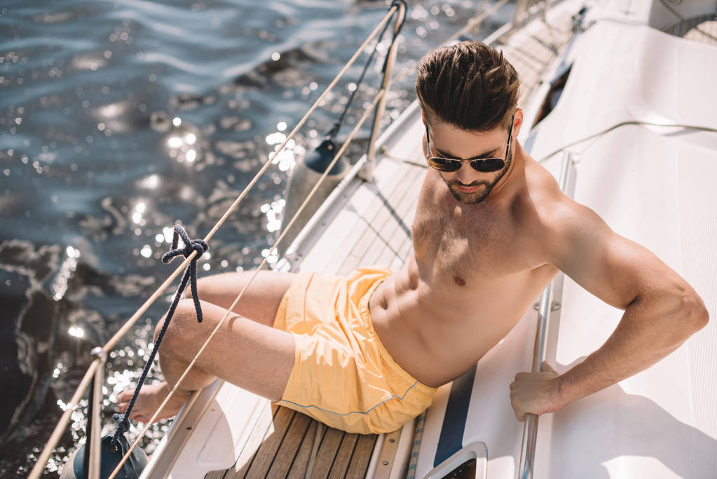 Мужчина без рубашки в плавках и солнцезащитных очках, загорающий на яхте
  - Фото, изображение