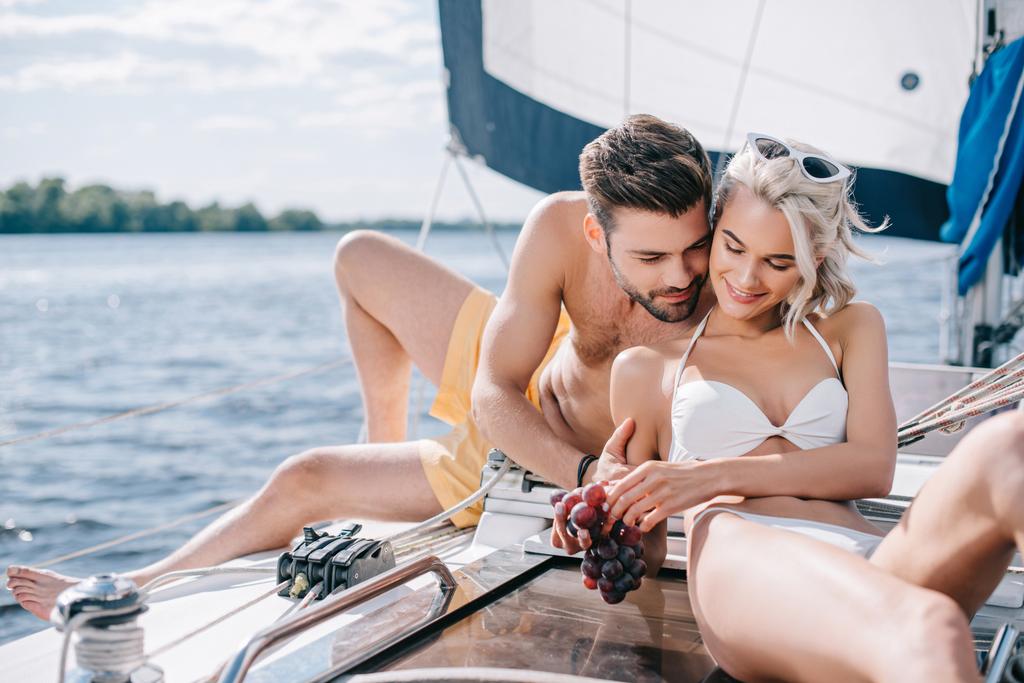 šťastný mladý pár v plavky uvolňující a jíst hrozny na jachtě  - Fotografie, Obrázek