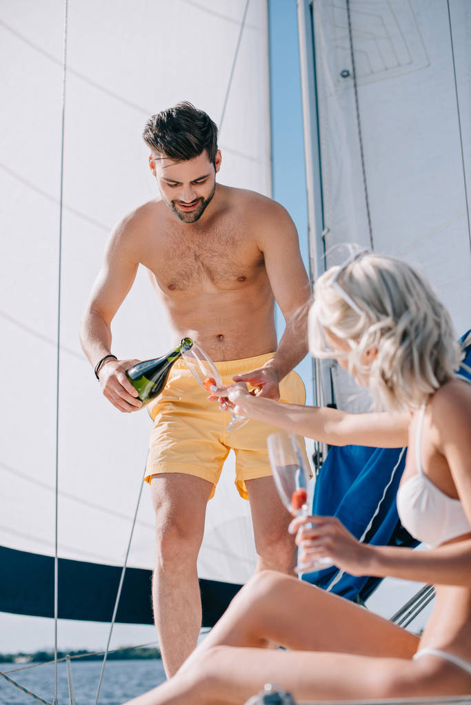 schöner hemdloser Mann in Badehose gießt Champagner in Glas von Freundin auf Jacht  - Foto, Bild