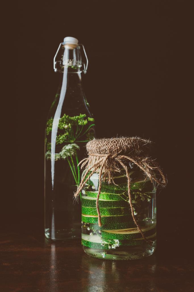 glass bottle with water and dill and glass jar with zucchini on wooden table on black - Photo, Image