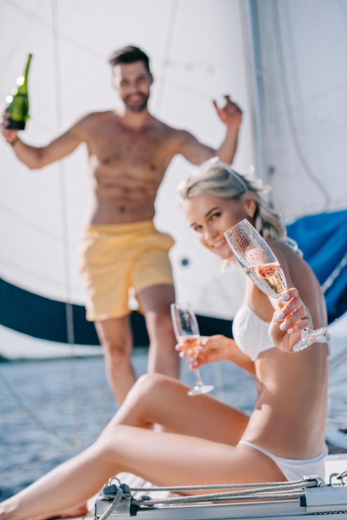 selective focus of smiling woman showing glass of champagne and her boyfriend standing behind on yacht  - Photo, Image