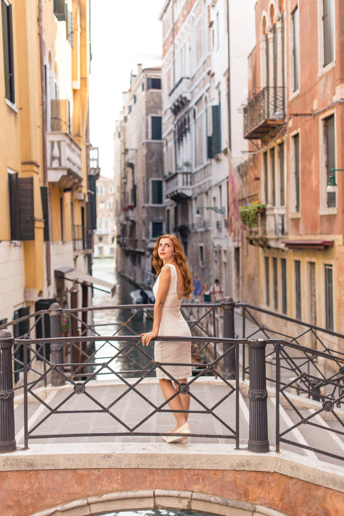 魅力的な赤毛の女性がヴェネツィアの狭い運河に橋のポーズ - 写真・画像