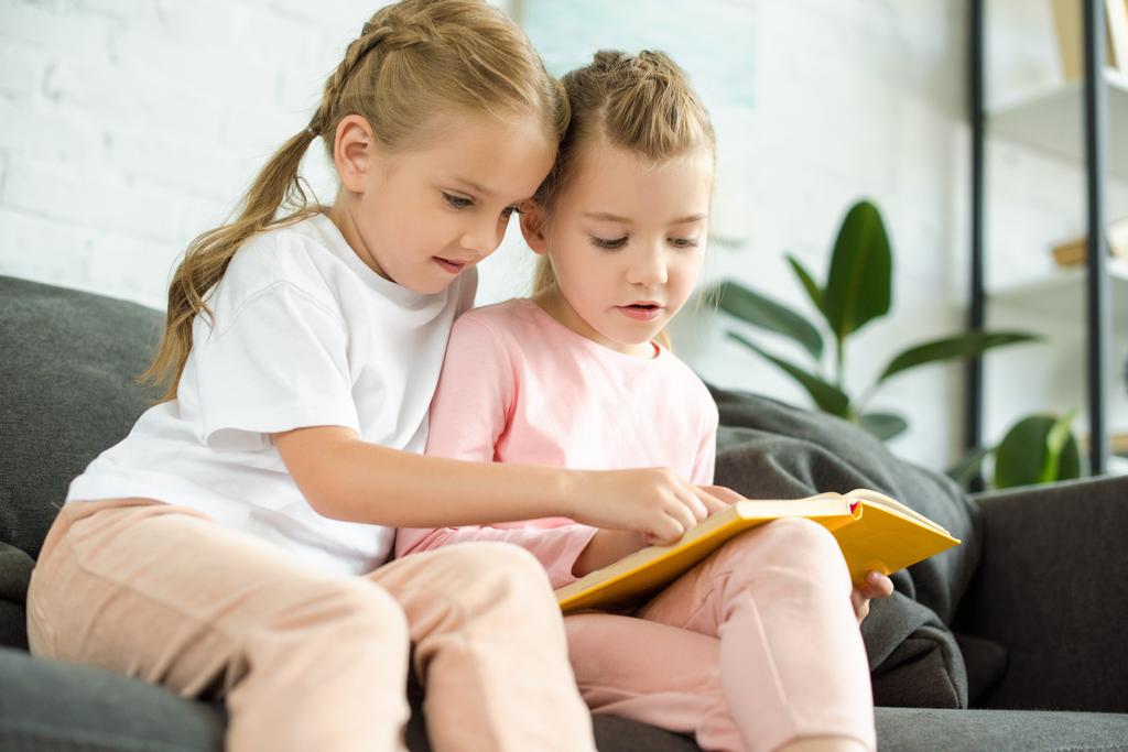 adorables hermanitas leyendo el libro juntas en el sofá en casa
 - Foto, imagen