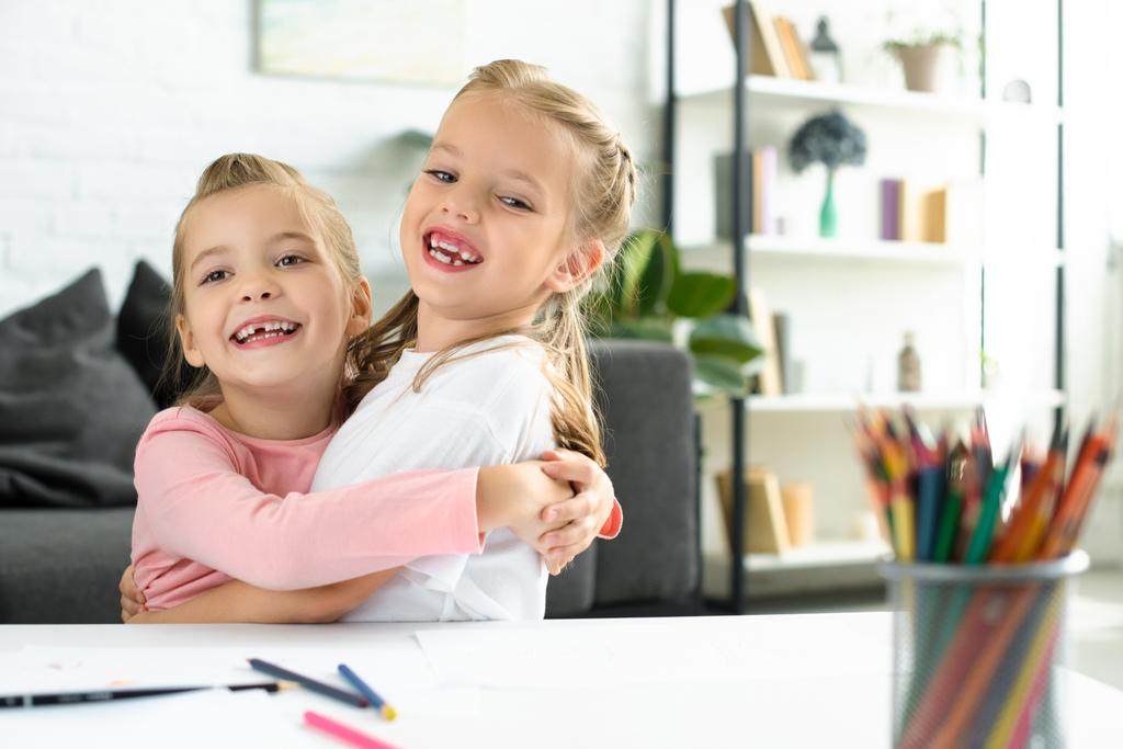 portrait of happy little children hugging each other at table with papers and pencils for drawing at home - Photo, Image
