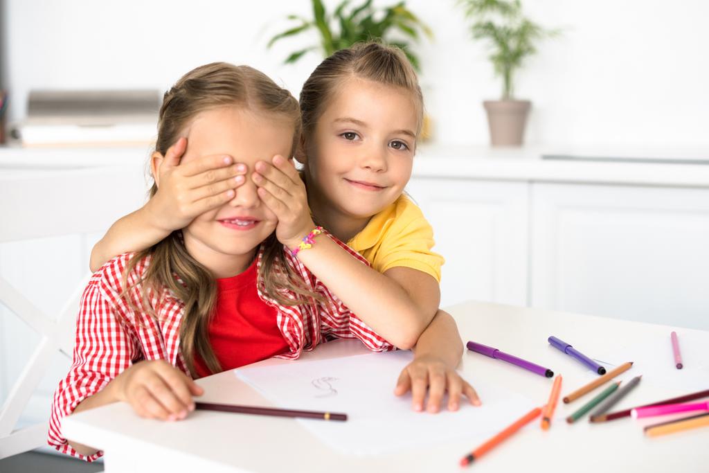 słodkie małe dziecko, obejmujących oczy, aby siostra przy stole z kredkami i papiery do rysowania w domu - Zdjęcie, obraz
