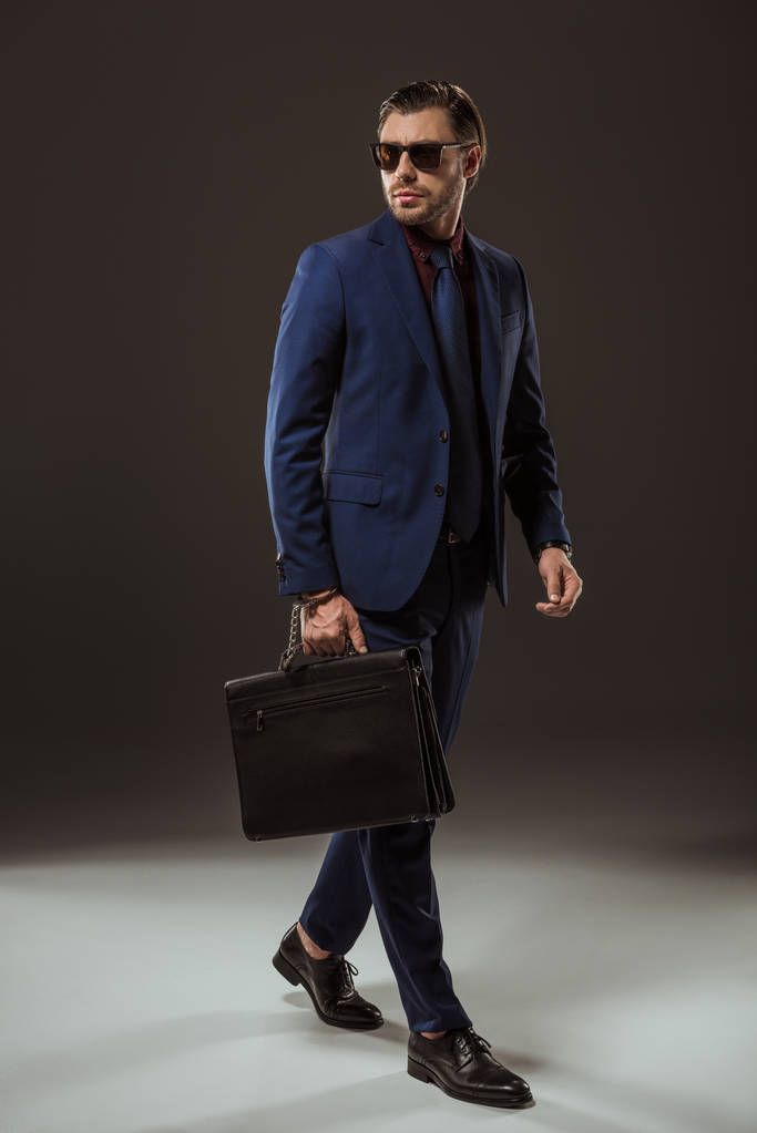 Вид в полный рост стильного бизнесмена в солнцезащитных очках, держащего в руках чехол и отводящего вдаль взгляд на черный
 - Фото, изображение