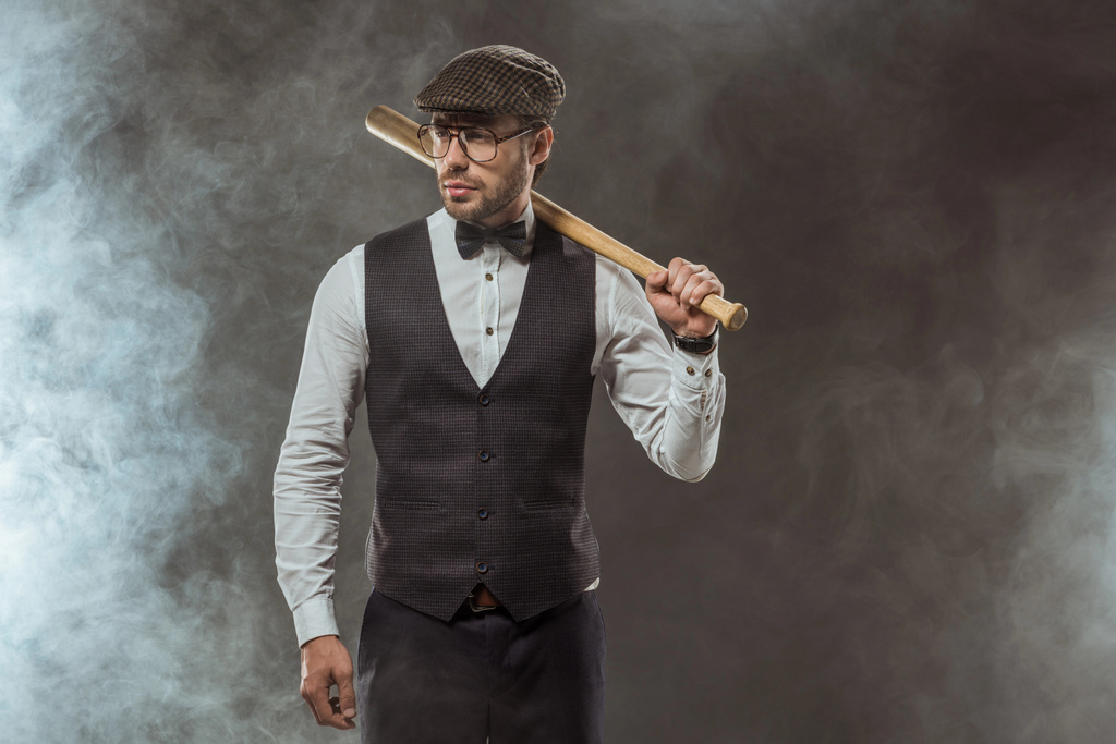 elegantní muž v motýlek, brýle a čepici držel baseballovou pálkou a koukal na černém - Fotografie, Obrázek