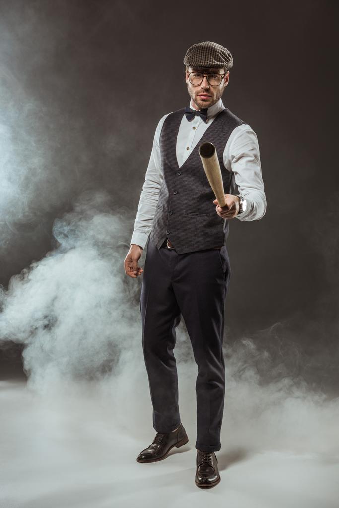 σοβαρή καλαίσθητο άνθρωπος παπιγιόν και καπάκι κρατώντας το ρόπαλο του μπέιζμπολ, ενώ στέκεται στον καπνό  - Φωτογραφία, εικόνα