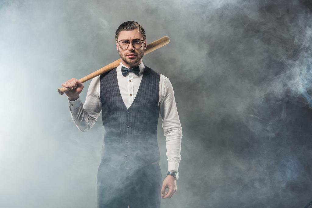 stylish man in bow tie and eyeglasses holding baseball bat and looking at camera in smoke  - Photo, Image