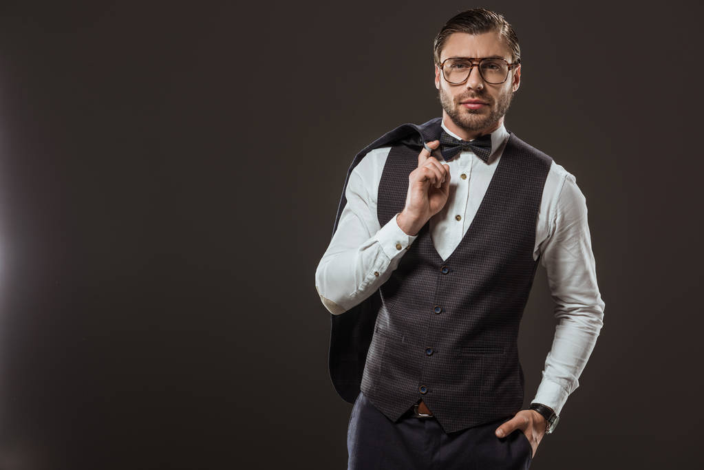 guapo hombre elegante en corbata de lazo y gafas con chaqueta de traje y mirando a la cámara aislada en negro
 - Foto, Imagen