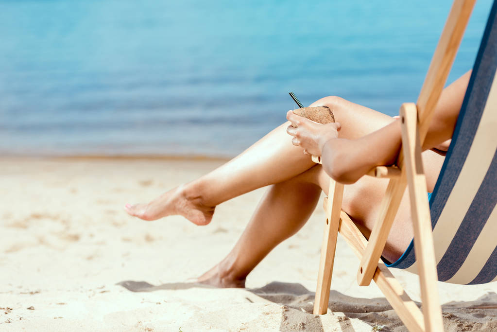 cropped image of woman holding cocktail in coconut shell and laying on deck chair on sandy beach  - Photo, Image