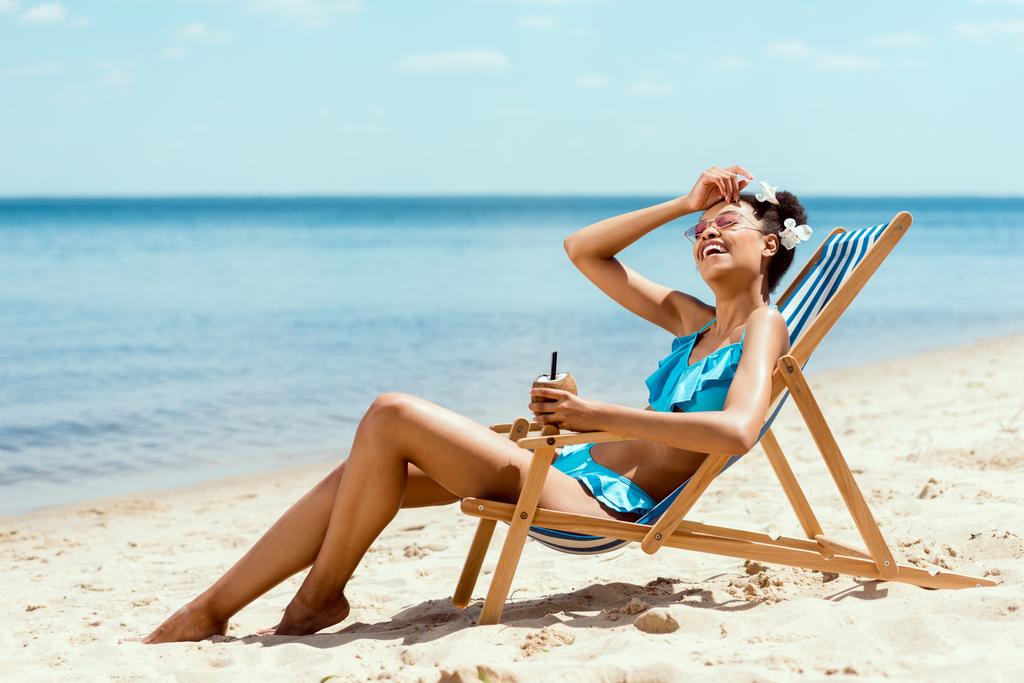 joven mujer afroamericana en bikini limpiando la frente y sosteniendo cóctel en cáscara de coco mientras está acostado en la silla de cubierta en la playa de arena
  - Foto, imagen