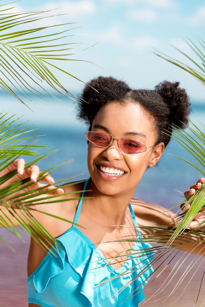 pozostawia uśmiechający się african american Kobieta w okulary w pobliżu palm nad brzegiem morza - Zdjęcie, obraz