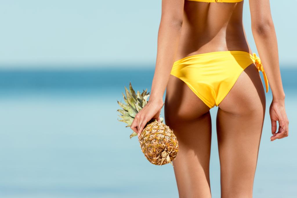 przycięty obraz kobiety w bikini, trzymając ananasa przed morza  - Zdjęcie, obraz