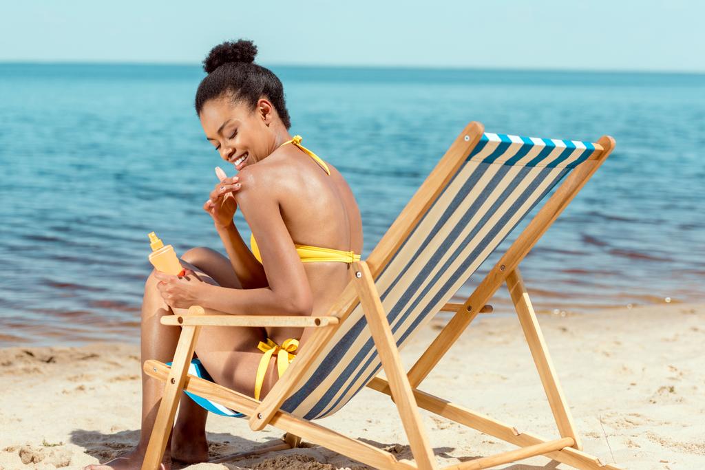 lächelnde Afroamerikanerin, die Sonnencreme auf die Haut aufträgt, während sie auf einem Liegestuhl am Sandstrand sitzt  - Foto, Bild