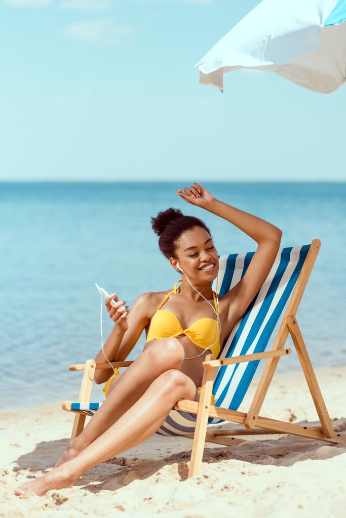 happy african american woman laying on deck chair and listening music in earphones with smartphone under beach umbrella in front of sea  - Photo, Image