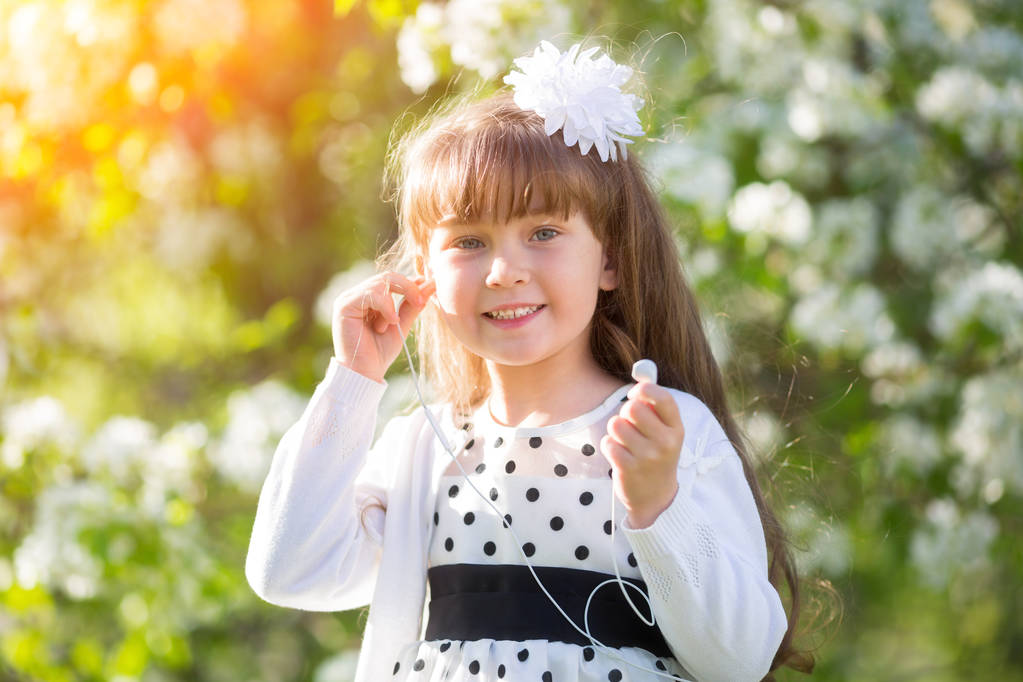 Маленькая девочка в белом платье слушает аудиозаписи в наушниках летом и в парке. Девушка слушает музыку со своего мобильного телефона
. - Фото, изображение