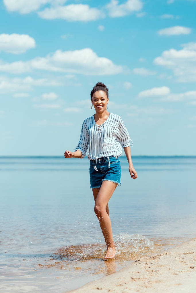 砂浜近くの海の水で走っているアフリカ系アメリカ人の女性の笑顔 - 写真・画像