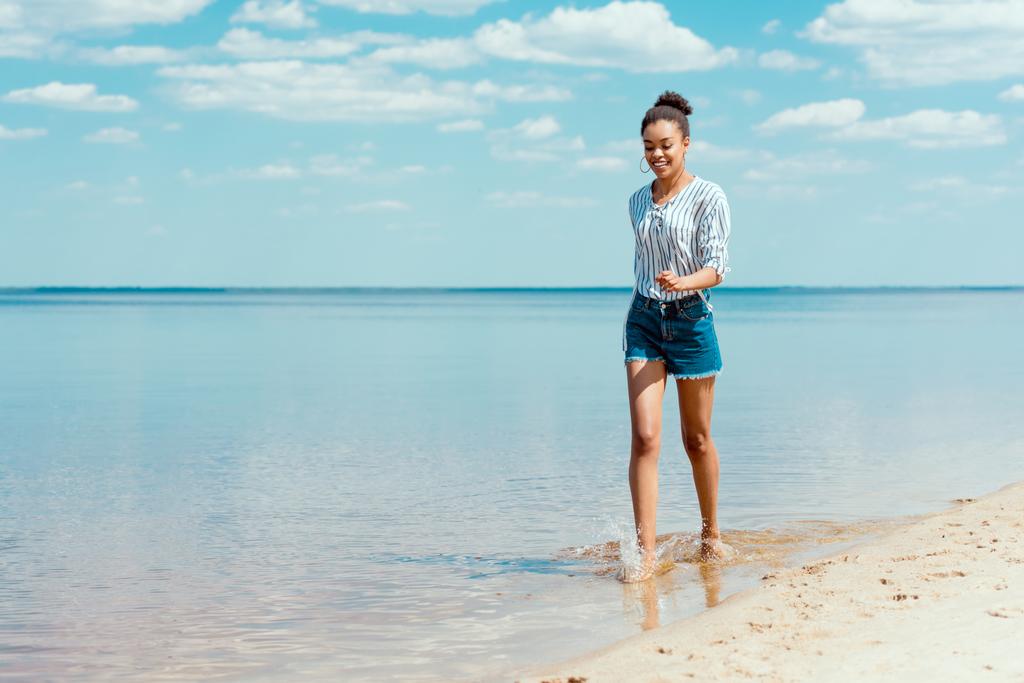 junge lächelnde Afroamerikanerin spaziert im Meerwasser in der Nähe von Sandstrand - Foto, Bild