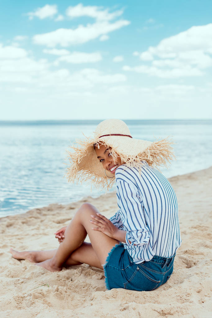 ευτυχισμένος αφρικανική αμερικανική γυναίκα σε ψάθινο καπέλο βλέπουν φωτογραφική μηχανή, ενώ κάθεται σε αμμώδη παραλία - Φωτογραφία, εικόνα