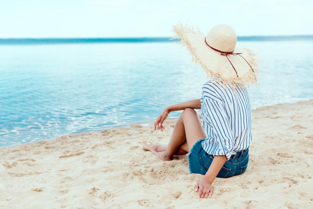 πίσω όψη του νεαρή γυναίκα στο χαλαρωτικό ψάθινο καπέλο στην παραλία  - Φωτογραφία, εικόνα