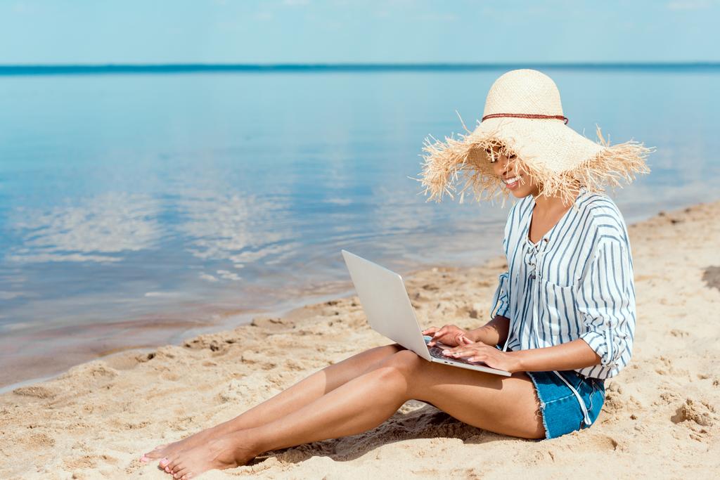 Happy africké americké ženské na volné noze v slaměný klobouk sedí na písečné pláži s notebookem  - Fotografie, Obrázek