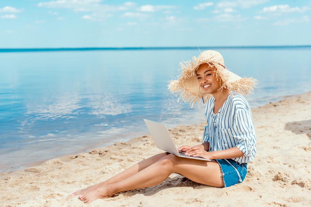 mosolyogva ül a laptop homokos strand szalmakalap-afrikai-amerikai női szabadúszó  - Fotó, kép