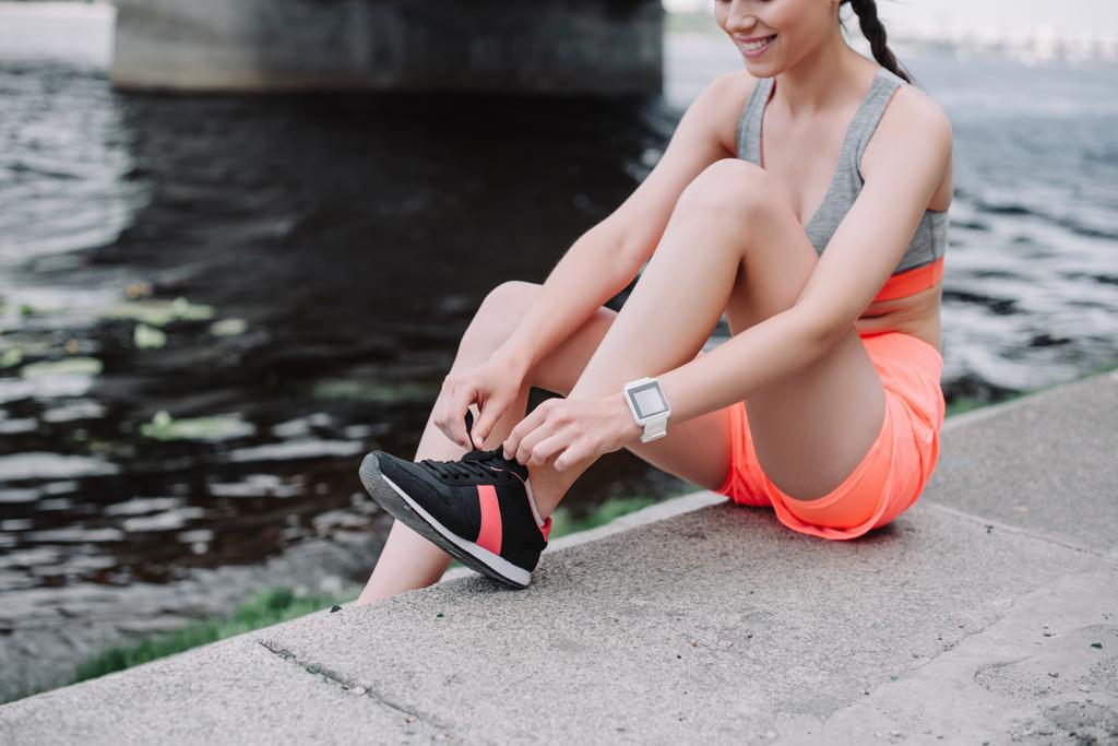 обрезанный вид спортсменки, завязывающей шнурки на кроссовки и сидящей на набережной
 - Фото, изображение