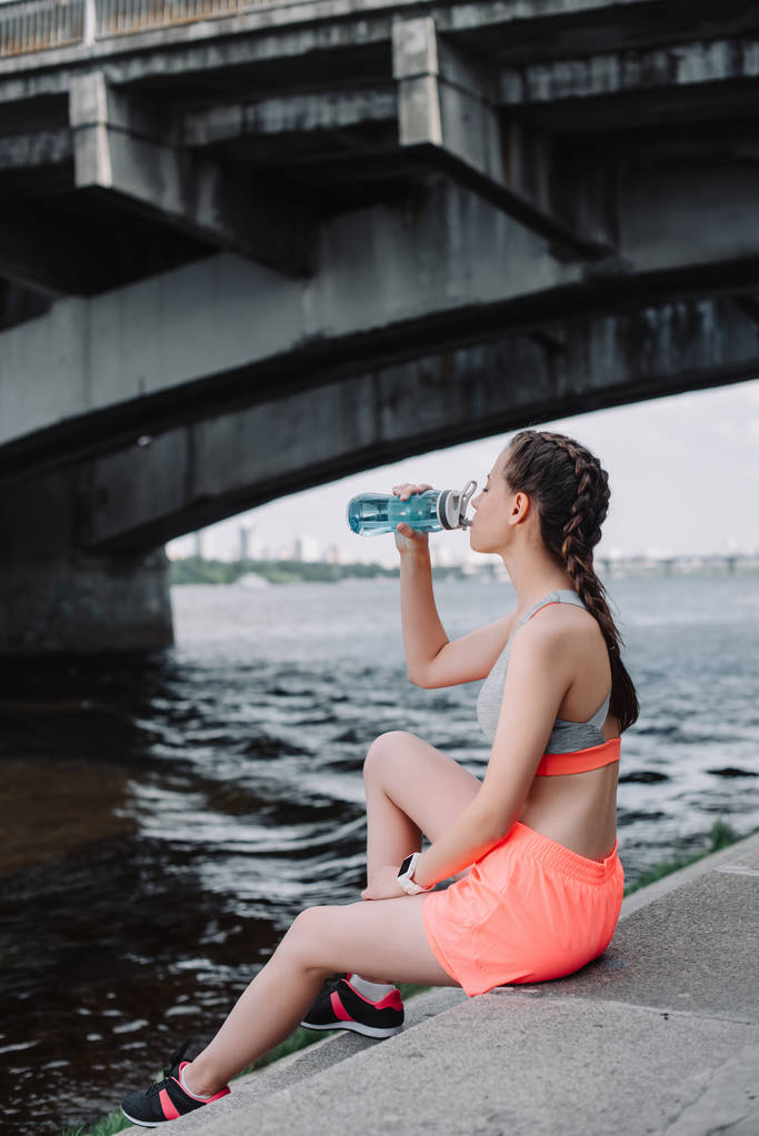 jovem atlético mulher beber água de esportes garrafa e sentado no cais perto do rio
 - Foto, Imagem
