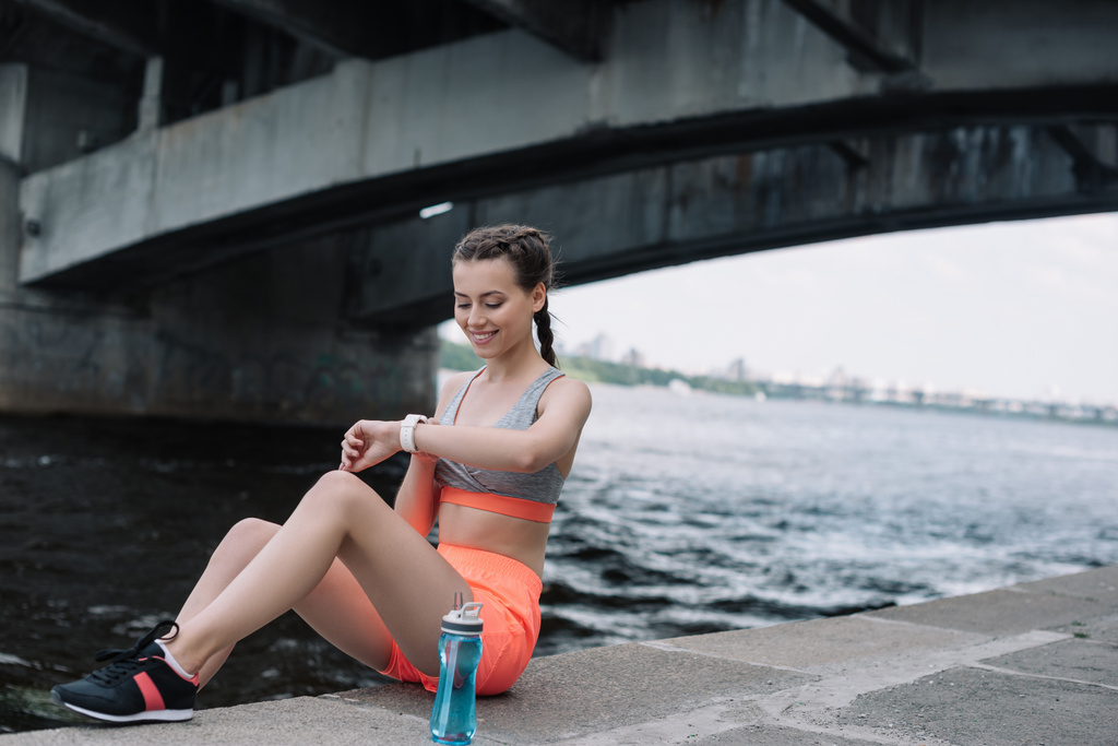 sorrindo esportista olhando para relógio inteligente enquanto sentado no cais com garrafa de água
  - Foto, Imagem