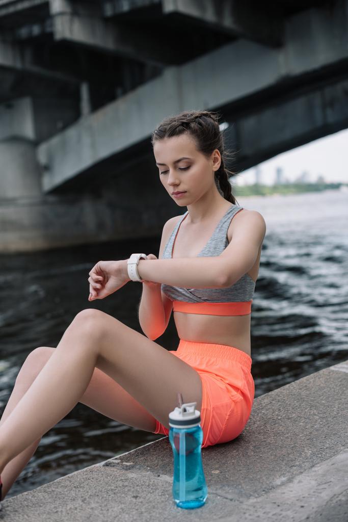 молодая спортсменка смотрит на умные часы, сидя на набережной с бутылкой воды
  - Фото, изображение