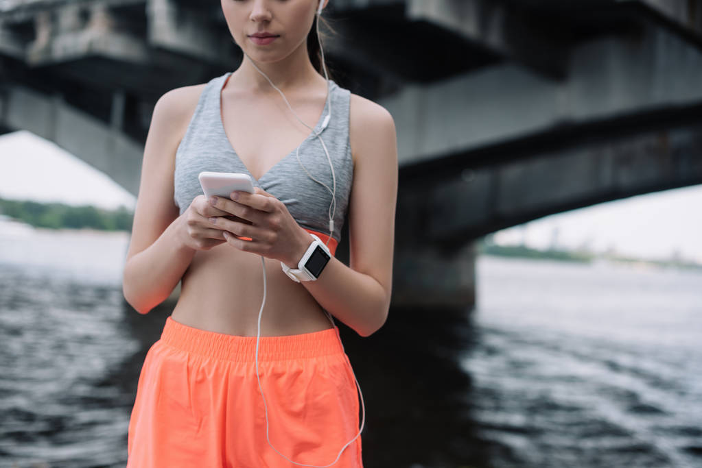 cropped view of young sportswoman using smartphone with earphones and smartwatch - Photo, Image
