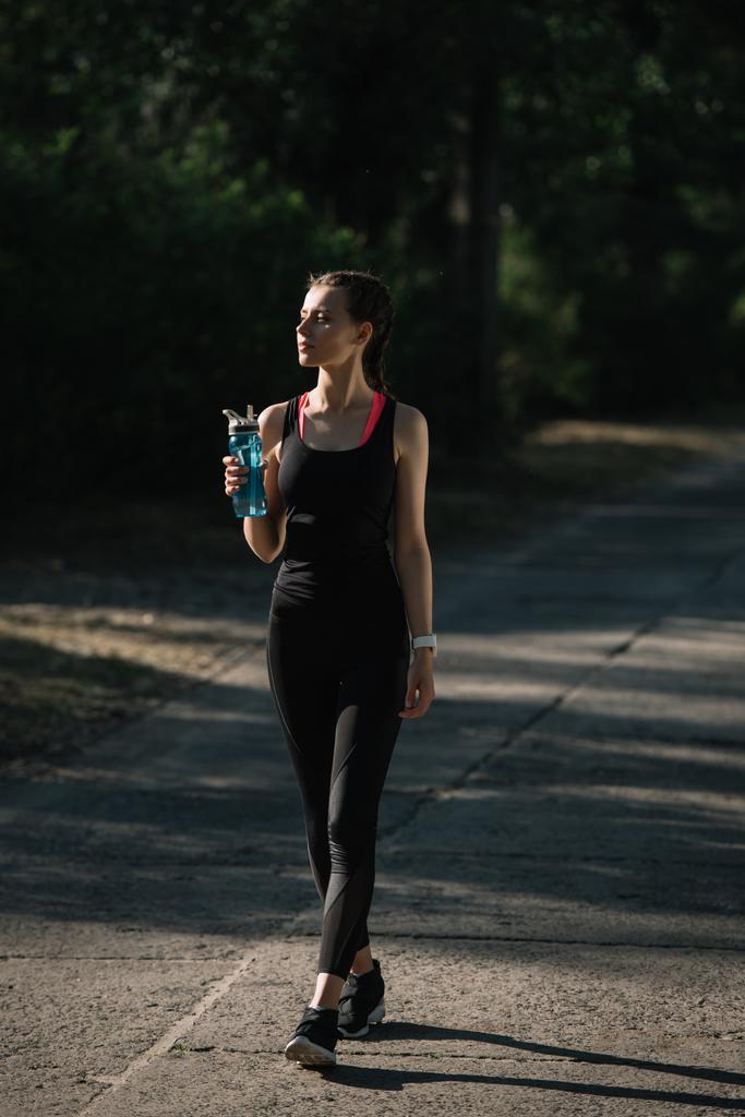 Sportlerin hält Sportflasche in der Hand und läuft auf Weg im Park - Foto, Bild