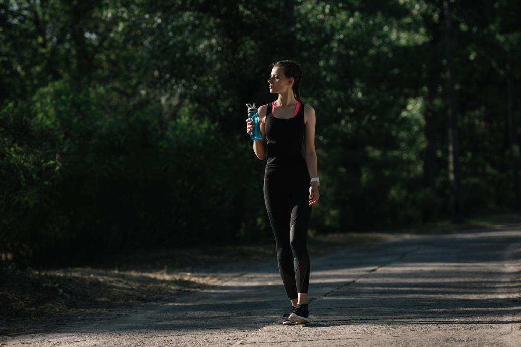 спортивна дівчина тримає спортивну пляшку з водою і ходить в парку
 - Фото, зображення