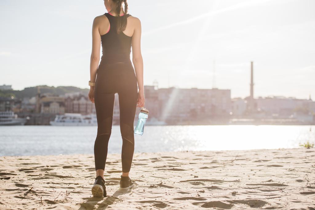 arkadan görünüşü atletik kadın spor şişe su ile arkadan aydınlatmalı sahilde yürüyüş ile - Fotoğraf, Görsel