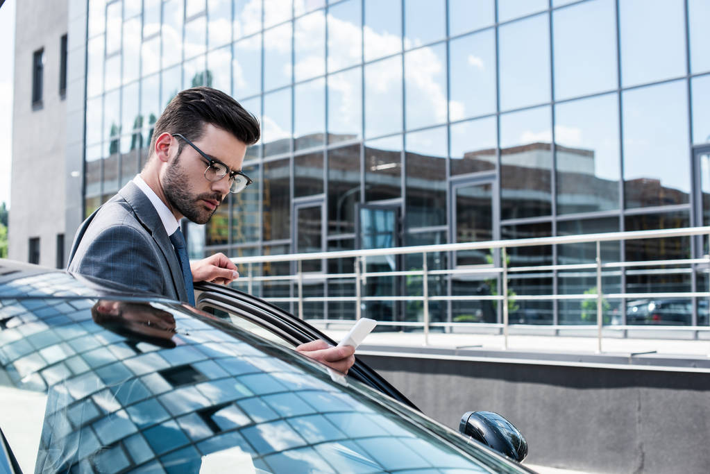 side view of focused businessman using smartphone while standing near car on street - Photo, Image