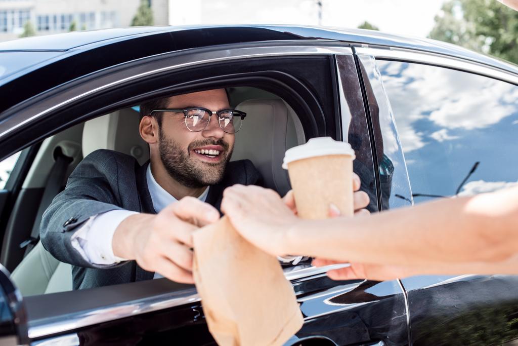 recortado disparo de sonriente hombre de negocios tomando café para ir y tomar la comida de camarero mientras está sentado en el coche
 - Foto, Imagen
